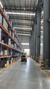Steel warehouse interior showing column spacing, pallet racks and an orange forklift in a marked aisle.